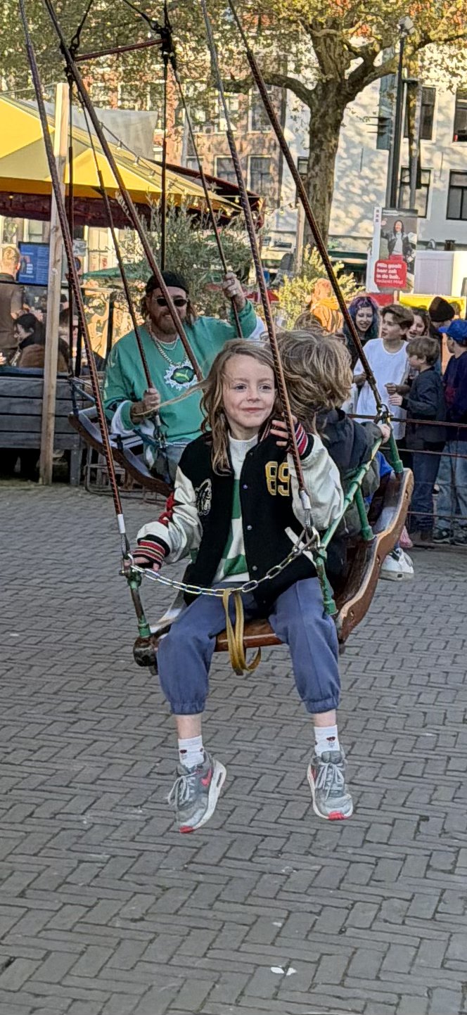 A group of people, including children and adults, are enjoying a carousel swing ride on a sunny day in Nieuwmarktbuurt, Amsterdam
